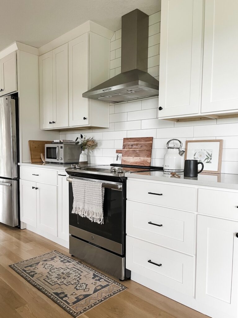 kitchen with white cabinets, light gray countertops and white backsplash tile. There is an oven and a vent plus kitchen accessories and decor. There is a rug in front of oven.
