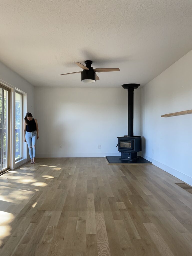 woman standing in empty room with white walls, white oak flooring, wood stove, and black flush mount ceiling fan