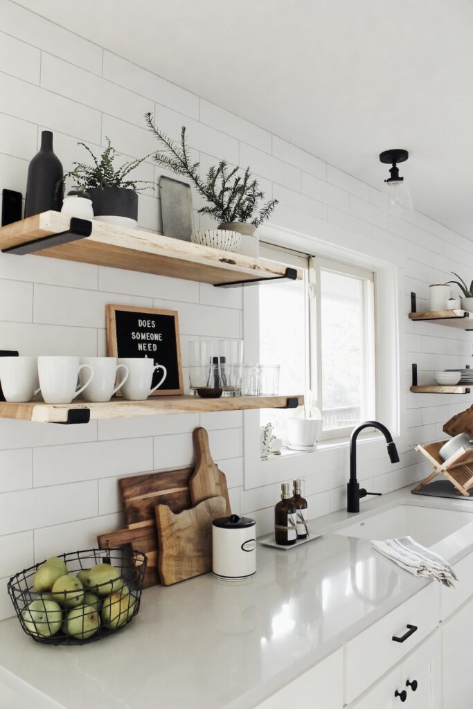 white kitchen with wood shelves and gray countertops. There are bowls, mugs, and vases on each shelf. There are cutting boards and a bowls of fruit on the counter.