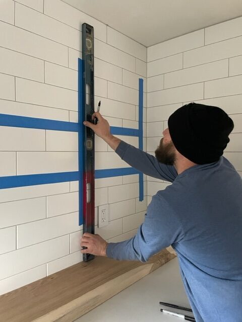 Man installing black shelf brackets on white subway tile by holding up a level. wood shelves on counter beneath
