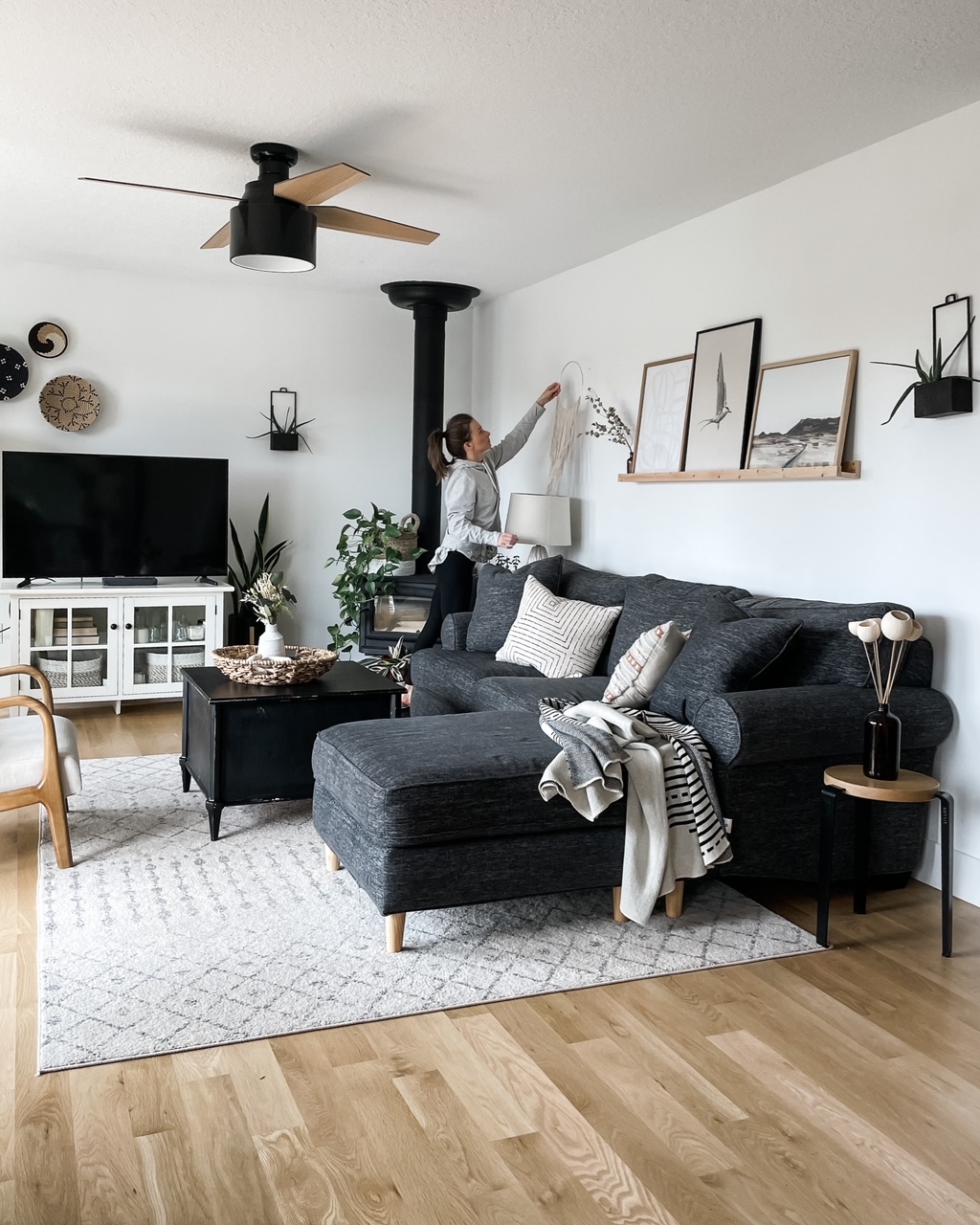 neutral family room with white walls, oak wood floors, dark gray couch, moroccan area rug, plants, and woman hanging up wall decor