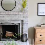 Brick fireplace white washed and round mirror above mantle, various decor items, including plants, artwork, and books. There is a vintage dresser nearby and text reads how to white wash a brick fireplace.