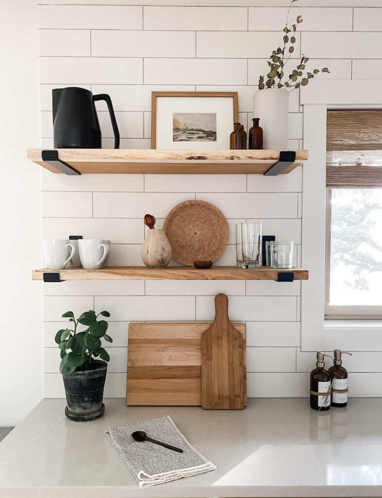 kitchen with two wood shelves and neutral decor including plants, frames, and cutting boards