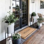 Front porch with double front doors painted with tricorn black and various decor items, including welcome at many plants grouped together in plant stands and planters, there are farmhouse style light fixtures, and wood deck. text below reads tricorn black Sherwin, Wiliams