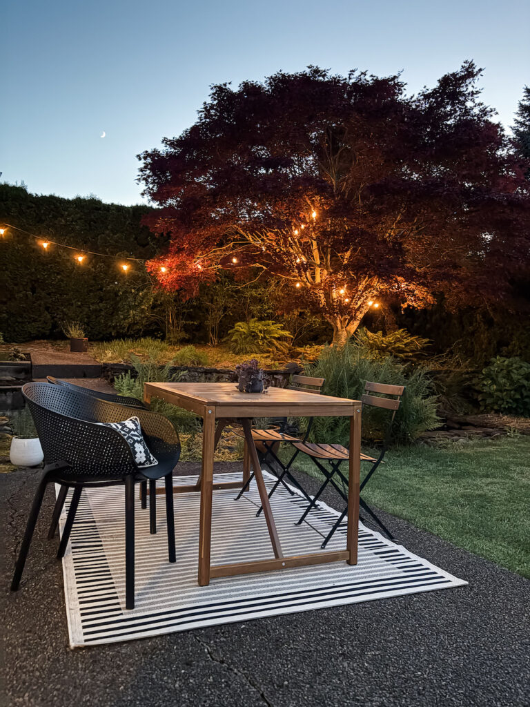 backyard patio with dining set on outdoor rug with chairs and twinkle lights in the trees.There is a lot of shrubs and plants