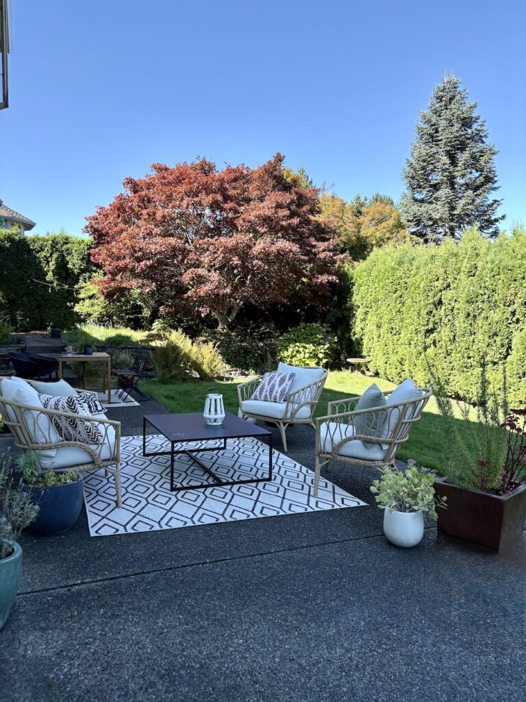 patio on concrete slab with outdoor rug, wicker furniture, and black coffee table. There are also throw pillows, potted plants, a lantern with grass and shrubs in the background.