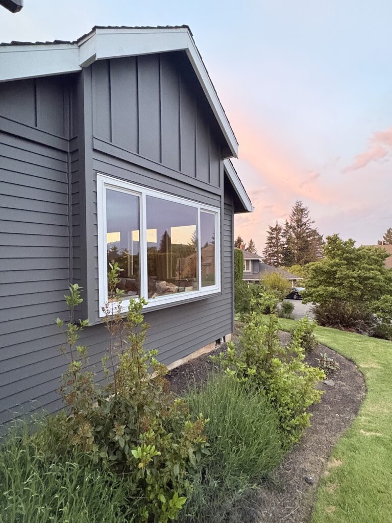 large window on dark gray house exterior with white trim and landscaping in front. There is a pinkish cloudy sky in  the background