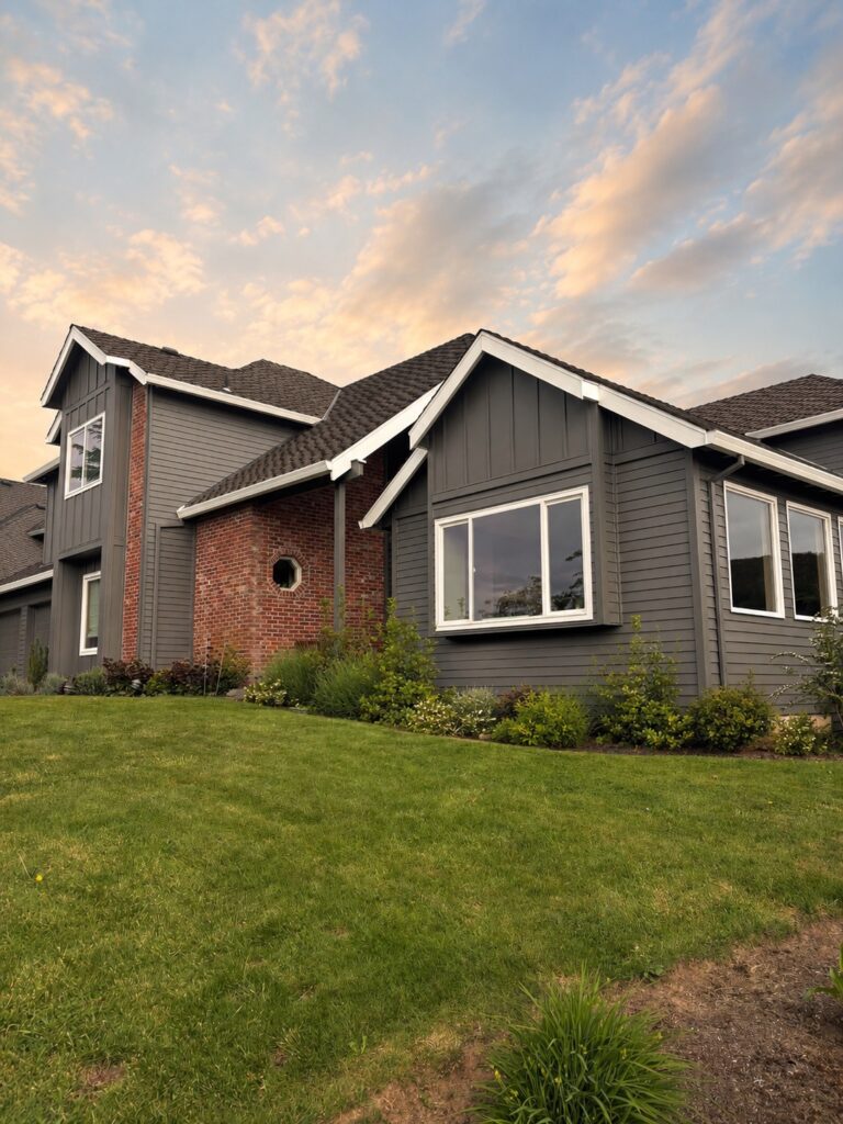 dark gray house exterior paint with white trim and brick details with lawn and landscaping. There is a pink blue sky in the background.