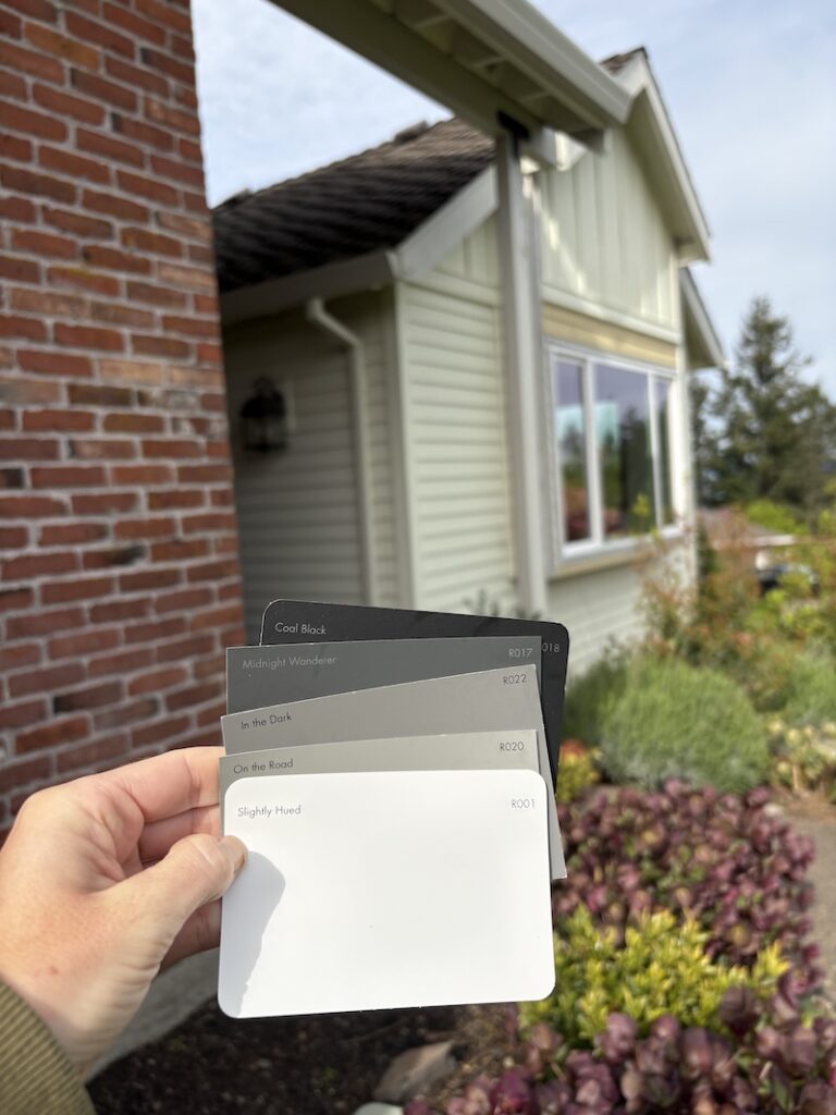 hand holding paint color chips including grays, white, and blacks. There is a creamy yellow house with brick in the background.