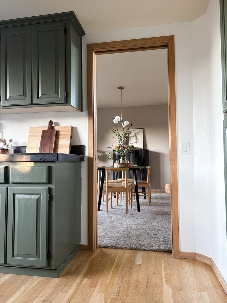 kitchen with rosemary green painted cabinets with wood trim and black countertops. There is cutting boards and a dining room in background.